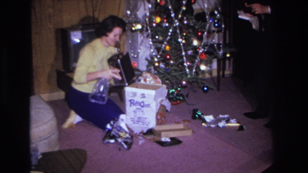 1966 home movie, Hoboken NJ: woman in yellow cardigan kneels on mauve carpet opening Christmas gift; tinsel-heavy tree and vintage CRT television visible in living room.