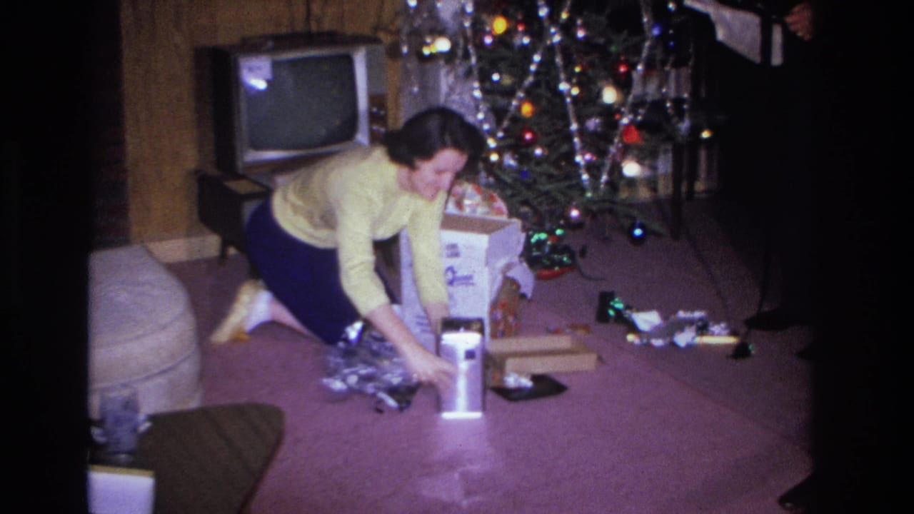 Woman in yellow sweater reaches toward an open gift box beside a tinsel-draped Christmas tree; vintage CRT television visible at right.