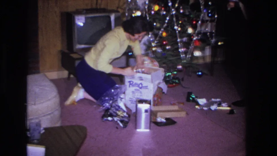 1966 home movie, Hoboken NJ: woman in yellow cardigan leans over opened gifts, branded product box with legible text and metallic tinsel spread across mauve carpet, Christmas tree behind.