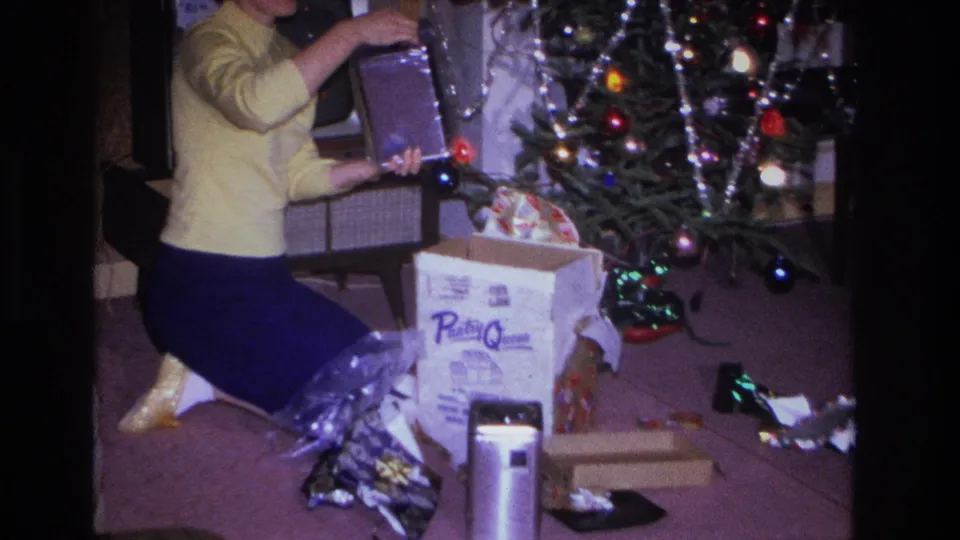 1966 home movie, Hoboken NJ: woman in yellow cardigan lifts a gift item from a branded product box beside a Christmas tree; vintage CRT television and wood-paneled walls visible.