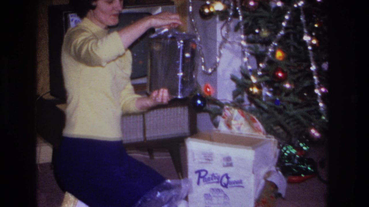 1966 home movie frame: woman in yellow sweater beside a lit Christmas tree in a Hoboken, New Jersey living room, with a 'Pantry Queen' branded carton on the floor.