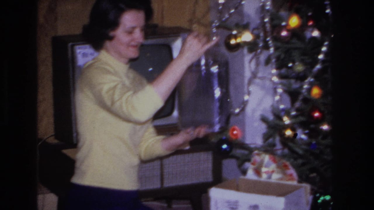Woman in yellow sweater reaches toward a lit Christmas tree; a mid-century television cabinet is visible in the background beside a branded carton.