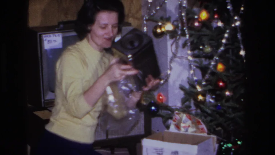 1966 home movie frame: woman unwrapping what appears to be a portable electronic device, possibly a tape player or radio, beside a lit Christmas tree.