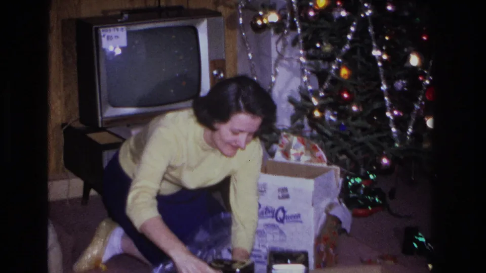 1966 home movie frame: woman crouching near gift boxes with a mid-century television and 'Pantry Queen' carton clearly visible beside a decorated tree in a New Jersey living room.