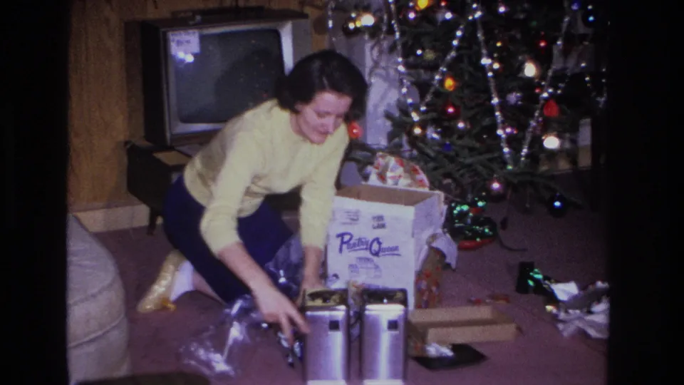 Mid-1960s Hoboken, NJ home movie showing a woman in a yellow sweater opening Christmas gifts beside a lit tree with tinsel and colored lights; CRT TV at left.