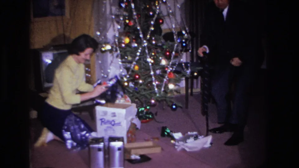 Mid-1960s home movie of a woman in a yellow sweater kneeling near a Christmas tree while a man in a dark suit stands in the background; branded box in foreground.