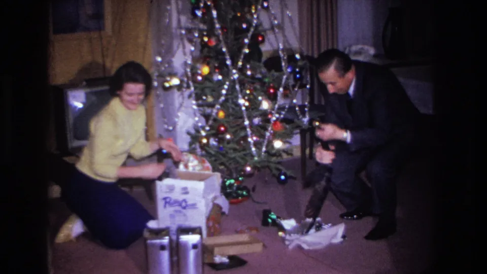 Mid-1960s Hoboken, NJ home movie of a man and woman examining a Christmas gift beside a lit tree with colored lights and tinsel; CRT TV visible at upper right.