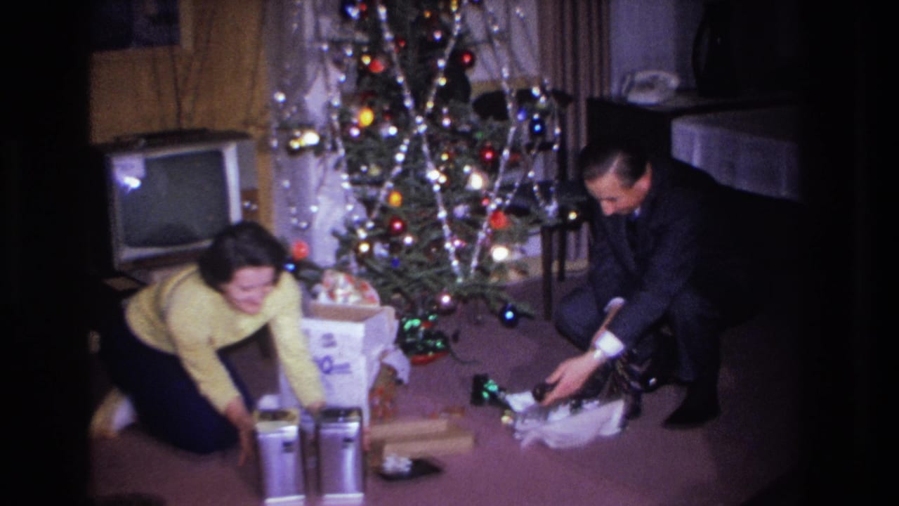 Woman in yellow sweater and man in dark suit opening Christmas gifts beside a lit tree in what appears to be a mid-1960s American living room; CRT television visible in background.