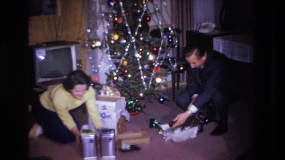 Woman in yellow sweater and man in dark suit opening Christmas gifts beside a lit tree in what appears to be a mid-1960s American living room; CRT television visible in background.