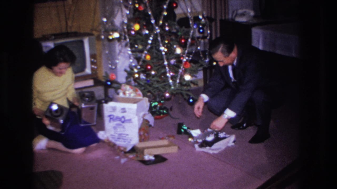Woman in yellow sweater and man in dark suit beside a Christmas tree with gifts spread across the floor.