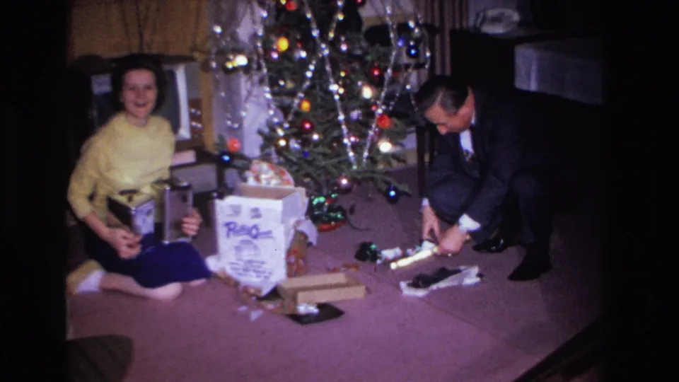 Woman in yellow sweater smiles while seated beside a lit Christmas tree; man in dark suit examines a gift in a mid-1960s domestic interior with a large branded gift box visible.