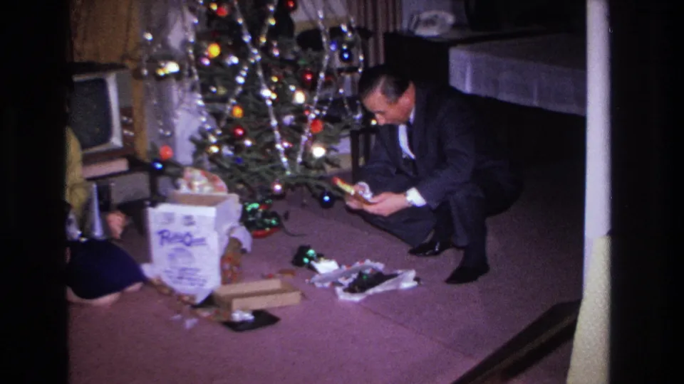 Man in dark suit crouches to examine a newly unwrapped item beside a decorated Christmas tree; large branded gift box and scattered packaging visible on purple carpet in a 1960s living room.