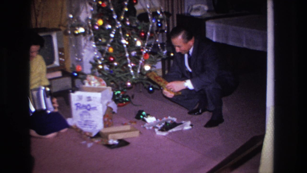 1966 Hoboken, NJ: man in dark suit and woman in yellow sweater open Christmas gifts on purple carpet; decorated tree with colored lights and partial figure at left.