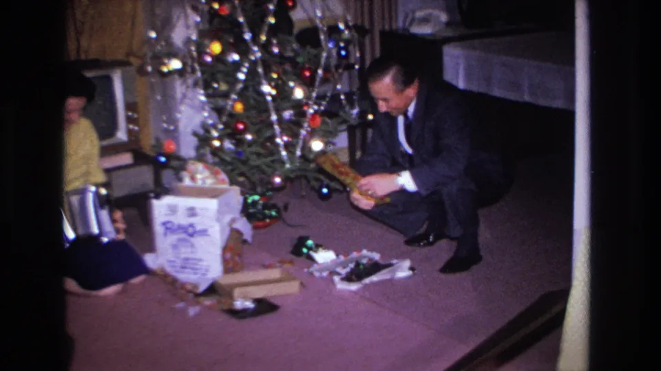 1966 Hoboken, NJ: man in dark suit and woman in yellow sweater open Christmas gifts on purple carpet; decorated tree with colored lights and partial figure at left.