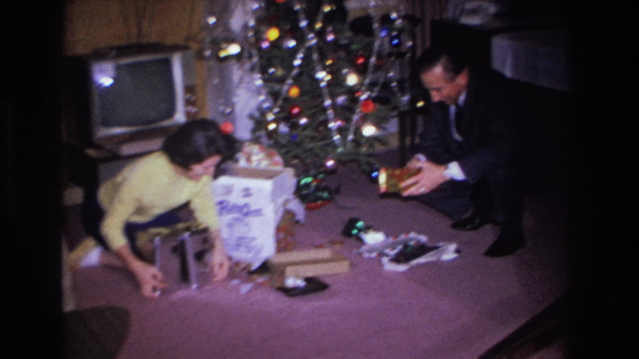 Woman in yellow sweater leans over gifts while man in dark suit reaches toward her; vintage CRT television visible beside the Christmas tree in background.