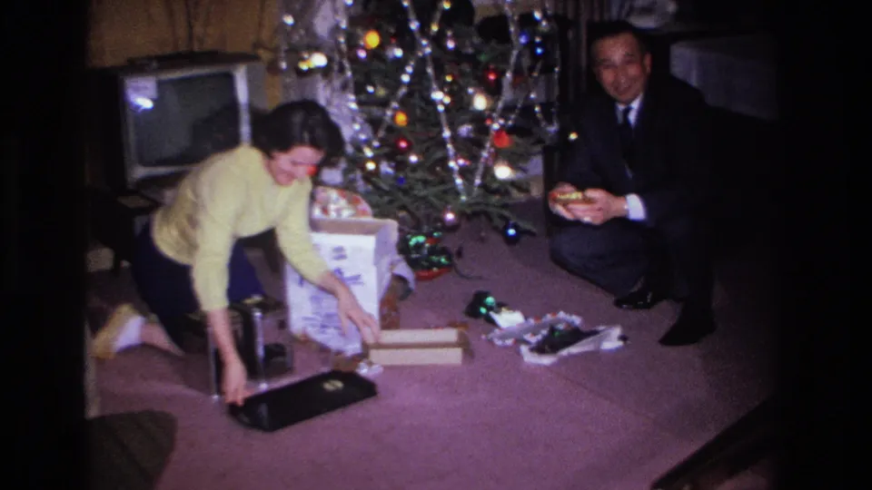 1966 Hoboken, NJ: woman reaches for an item on the floor as man holds a newly unwrapped gift; decorated tree and vintage television in background.