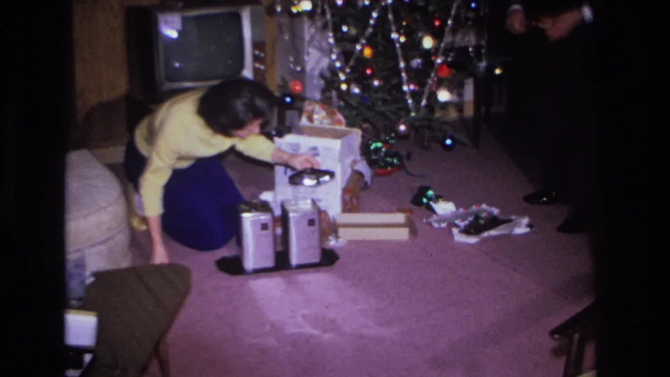 1966 Hoboken, NJ: woman examines a small portable electronic device among Christmas gifts; vintage CRT television and decorated tree in background.
