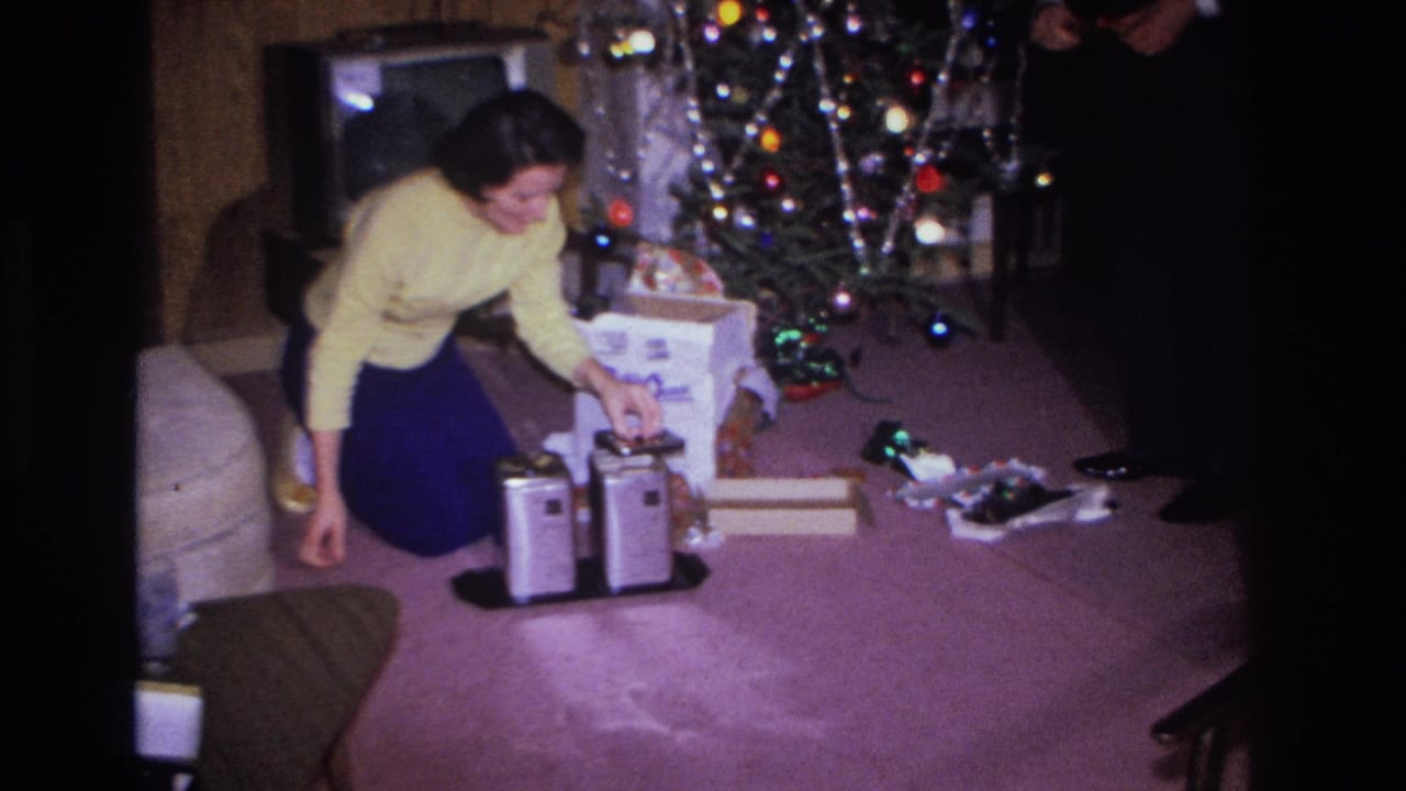1966 8mm home movie still: woman in yellow sweater kneeling over an open gift box near a chrome appliance, decorated Christmas tree with multicolored lights behind her, Hoboken NJ.