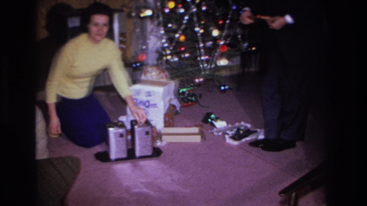 Woman in yellow sweater smiling near open gifts and chrome appliance; man in dark suit stands at right edge, partially out of frame.