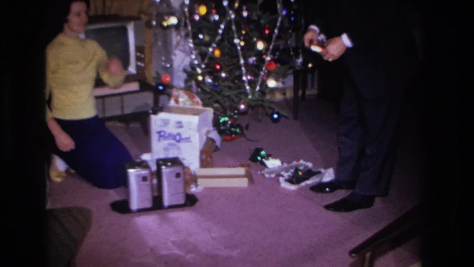 1966 8mm home movie still, wider framing: woman in yellow sweater turns toward left edge where a possible additional figure appears; man in dark suit holds object; decorated tree and opened boxes on carpet, Hoboken NJ.
