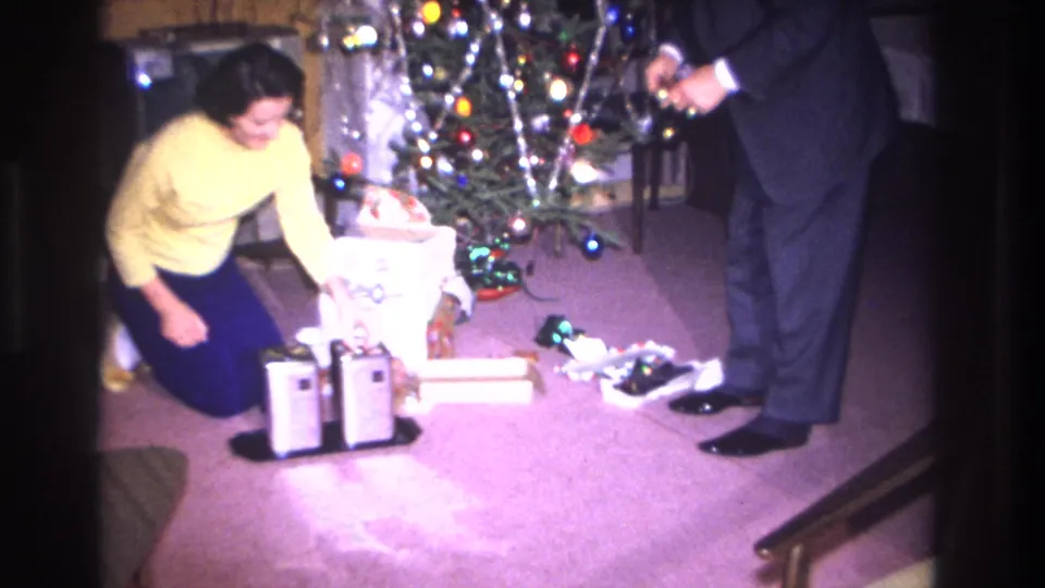1966 8mm home movie still: woman in yellow sweater crouching to unwrap a gift bag at the Christmas tree; man in dark suit at right, chrome appliance and scattered paper on mauve carpet, Hoboken NJ.