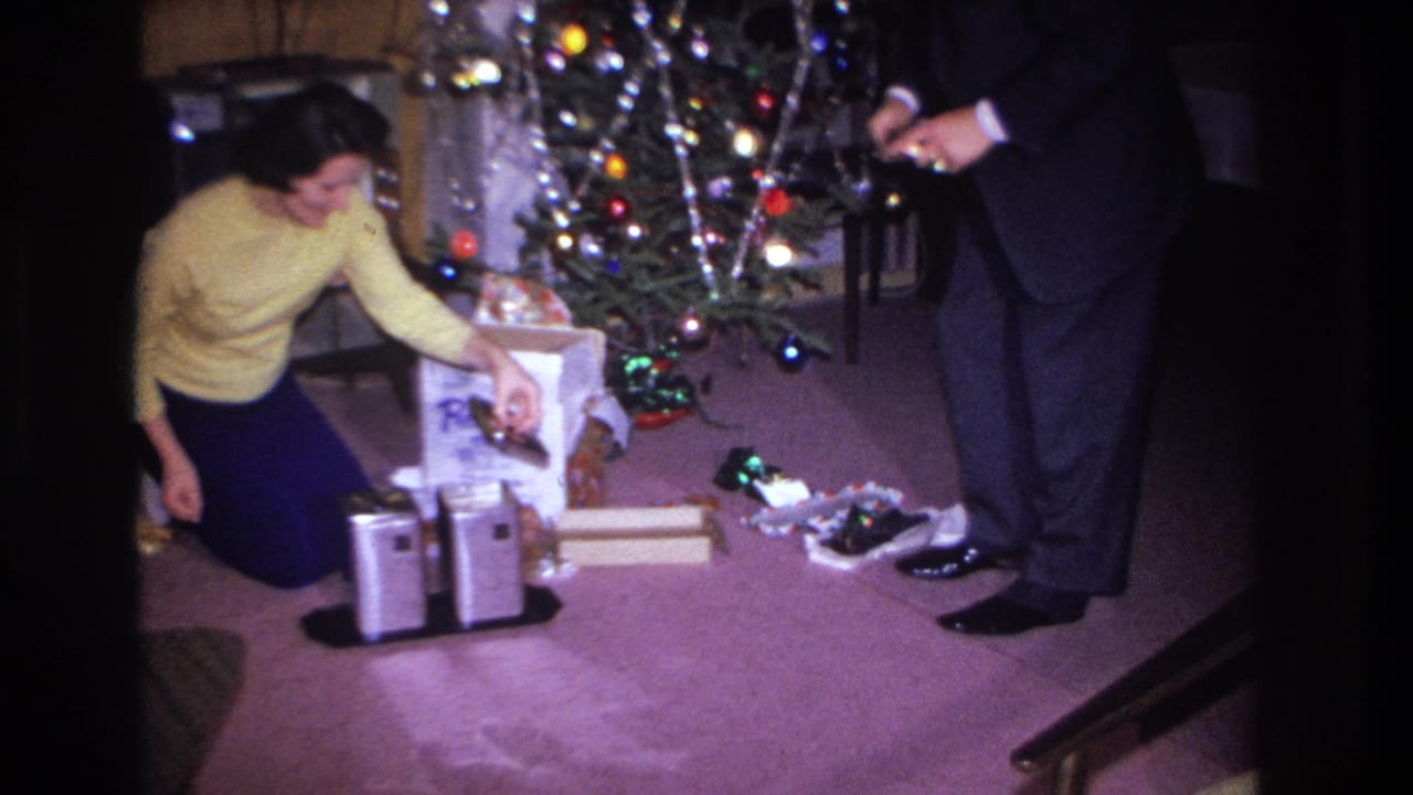 Woman in yellow sweater crouching beside a decorated Christmas tree with colored ball ornaments; man in dark suit standing nearby; 8mm home footage dated 1966, Hoboken NJ.