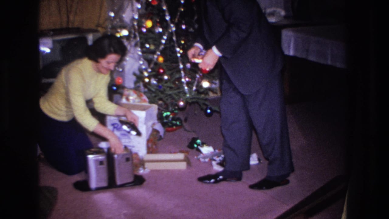 Woman in yellow sweater reaching toward a white product box near a lit Christmas tree; man in dark suit standing at right, hands raised.