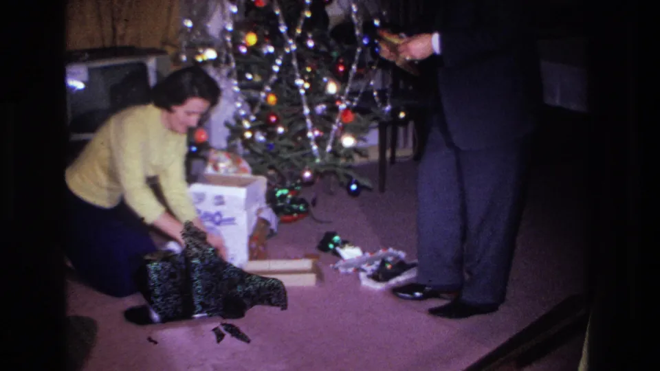 Woman in yellow sweater lifts what appears to be a dark garment from a gift box beside a decorated Christmas tree; 8mm home footage circa 1966, Hoboken NJ.