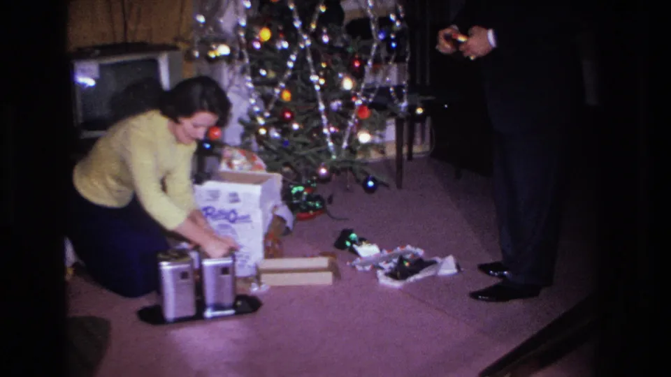 Woman in yellow sweater sorts through gifts on a pink carpet beside a decorated Christmas tree; 8mm home footage circa 1966, Hoboken NJ.