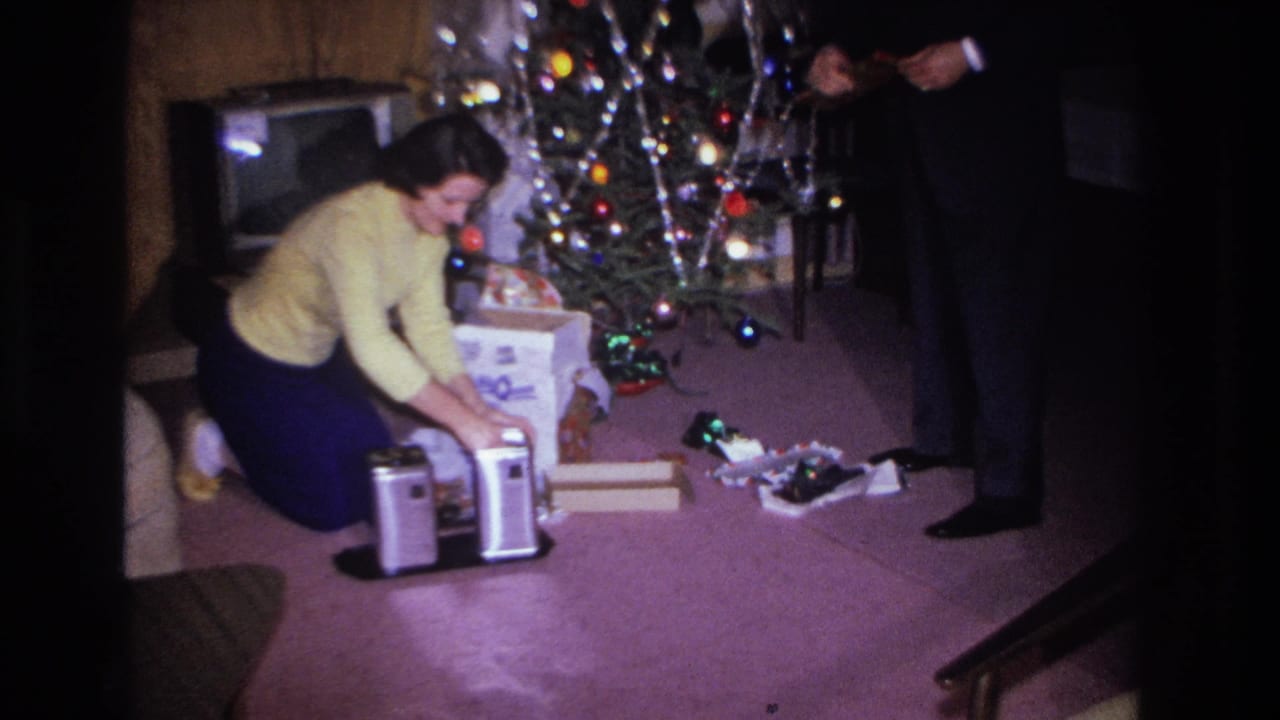 Woman in yellow sweater opening gifts beside a tinsel-and-bulb-lit Christmas tree in what appears to be a 1966 Hoboken, New Jersey living room, man in dark suit standing nearby.