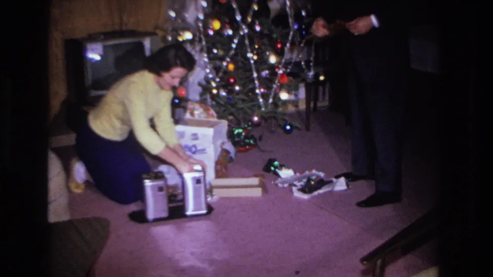 Woman in yellow sweater opening gifts beside a tinsel-and-bulb-lit Christmas tree in what appears to be a 1966 Hoboken, New Jersey living room, man in dark suit standing nearby.