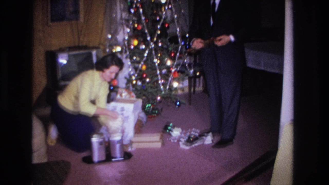 Woman in yellow sweater kneels over gifts at base of lit Christmas tree; man in dark suit stands to her right; vintage television visible at rear.
