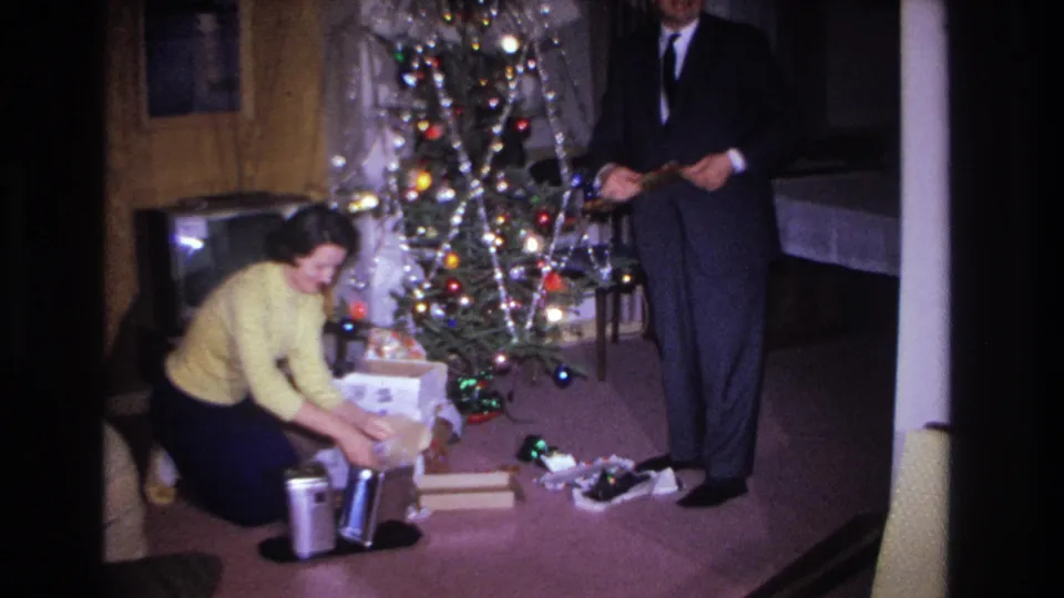 Mid-action frame of a woman sorting gifts beneath a tinsel-and-light-decorated tree as a man holds a small item, consistent with a 1960s American holiday home movie.