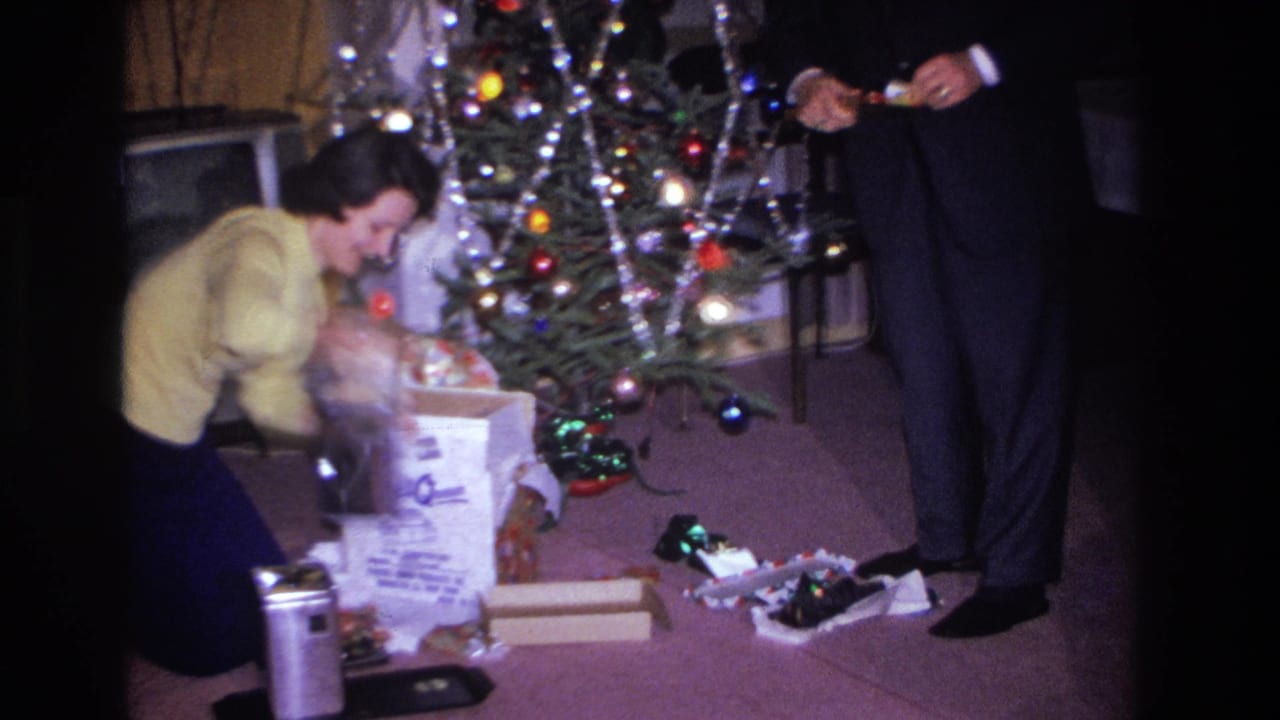Woman in yellow sweater opening Christmas gifts beside a decorated tree in a mid-century living room, Hoboken, New Jersey, circa 1966; man in dark trousers visible at right.