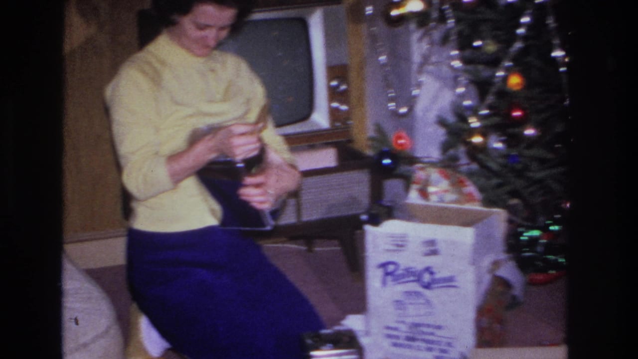Woman examines an unwrapped gift in a mid-century living room with a wood-cabinet television, Hoboken, New Jersey, circa 1966; white product box with printed text visible at right.