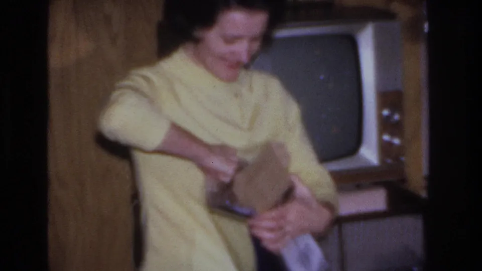 Woman in yellow sweater smiling while unwrapping a Christmas gift in a mid-century living room, circa 1966, Hoboken, New Jersey; CRT television visible in background.