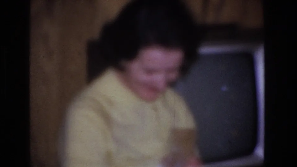 Out-of-focus close-up of woman in yellow sweater looking down in a mid-century living room, circa 1966, Hoboken, New Jersey; retro television visible in background.