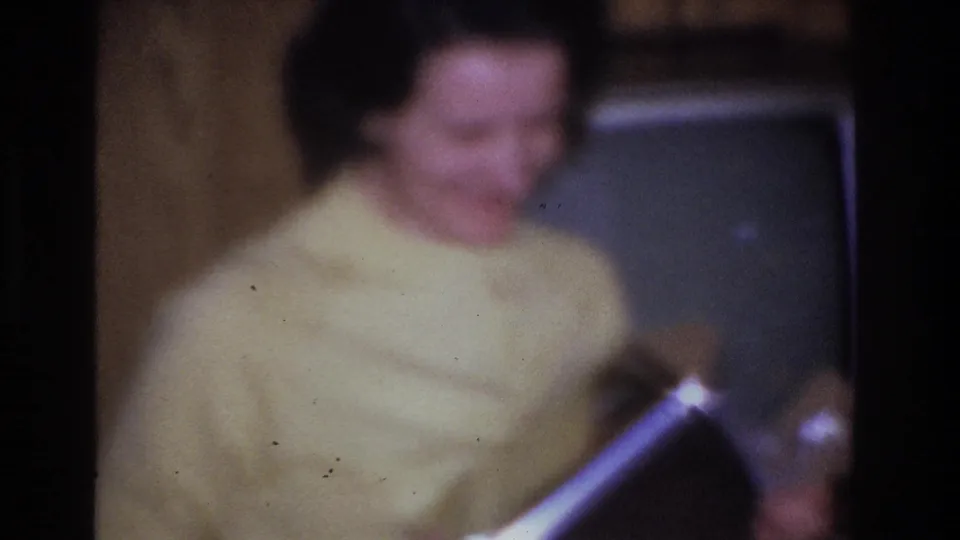Blurred close-up of a woman in a yellow sweater in what appears to be a mid-1960s American living room; a boxy vintage television is partially visible behind her.