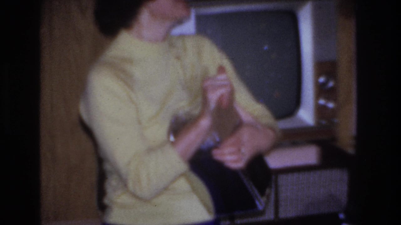 Woman in yellow sweater and blue trousers holding and inspecting an item in a 1960s living room, smiling; a boxy period television is visible behind her.