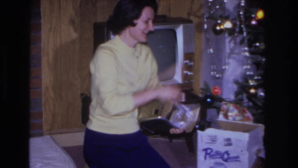Woman in yellow sweater and blue trousers opening a cardboard gift box in a 1960s American living room; a decorated Christmas tree with colored lights and a vintage TV are visible behind her.