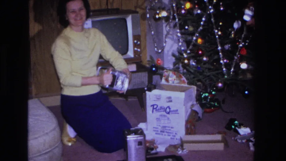 Woman in yellow sweater and blue trousers holding a wrapped gift beside a lit Christmas tree in a 1966 New Jersey living room; a branded product box is visible on the carpet.