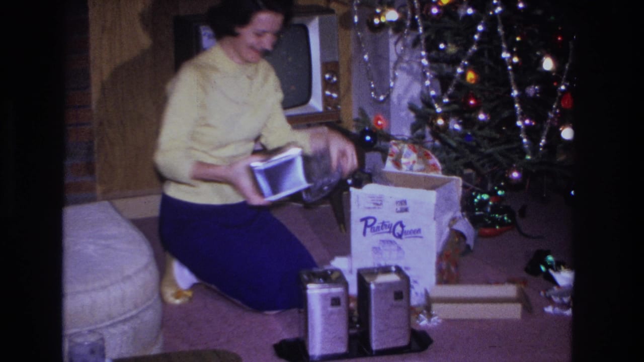 In a 1966 Hoboken home interior, a woman in a yellow sweater kneels by a decorated Christmas tree with gifts and metal canisters nearby.