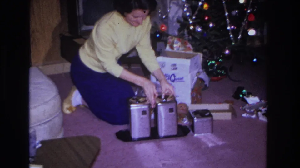 A woman kneels on the carpet and adjusts metal canisters beside an open box and decorated Christmas tree in a private holiday interior.