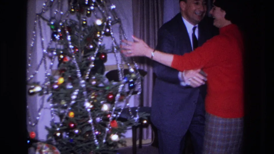A man in a suit and a woman in a red top appear to dance or embrace near the Christmas tree in the 1966 Hoboken home-movie scene.