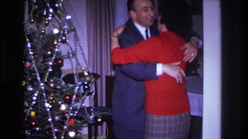 In 1966 Hoboken home-movie footage, a suited man embraces a woman in a red sweater near a Christmas tree.