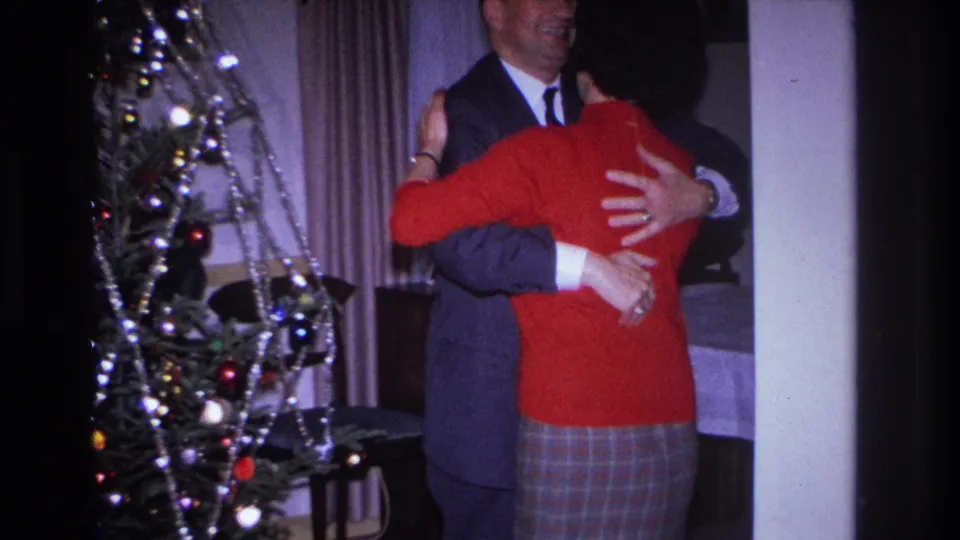 The woman in a red sweater faces away while embracing a suited man near a decorated tree in the Hoboken interior.
