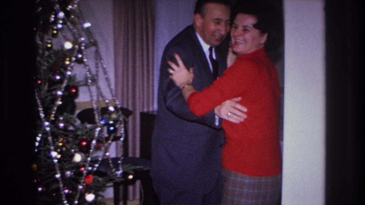 Man and woman face the camera while embracing, both smiling, beside a Christmas tree with colored bulb ornaments.