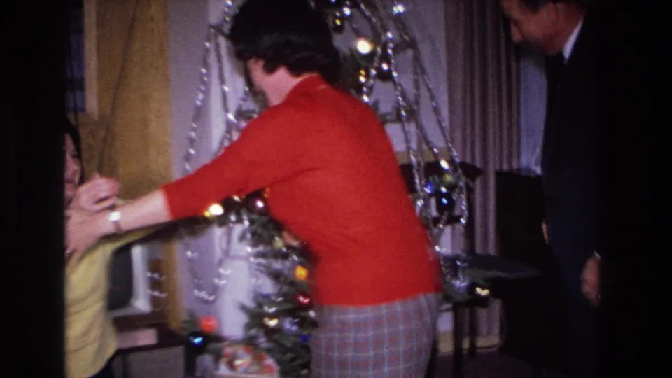 Woman in red sweater and plaid skirt near tinsel-decorated Christmas tree, with a shorter figure and a partial male figure at the right edge, Hoboken, New Jersey, circa 1966.
