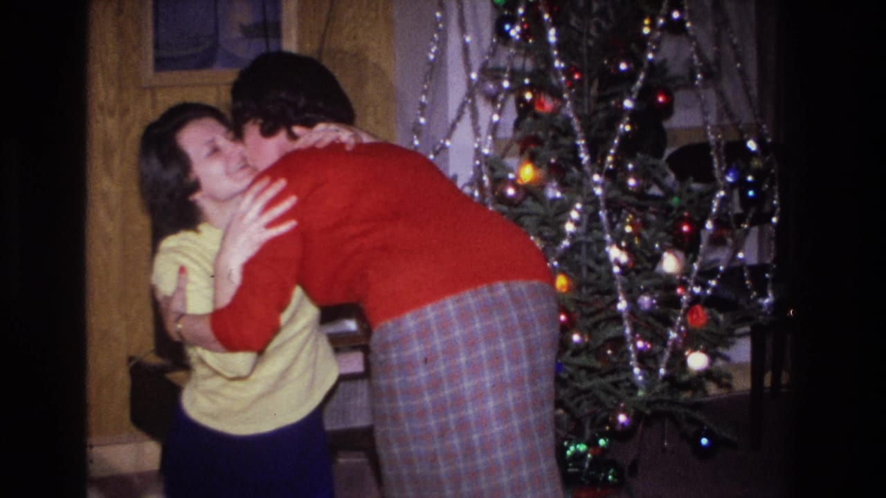 Two women hug in front of a tinsel-and-light-decorated Christmas tree in what appears to be a mid-1960s American home; one wears a yellow sweater, one a red sweater and plaid skirt.
