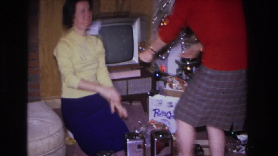 Two women crouch to examine gifts beside a mid-1960s Christmas tree; a boxy period television and a cardboard gift box with partial brand text are visible behind them.